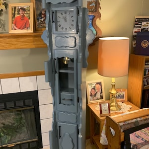 May include: A tall, gray painted grandfather clock with a decorative top and Roman numeral clock face. The clock has a glass door and several small drawers. The clock is standing in a room with a fireplace and a lamp.