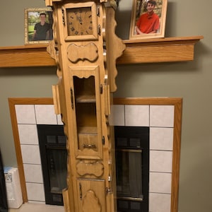 May include: A tall, light-colored wooden grandfather clock with a square clock face featuring Roman numerals. The clock has a cabinet with glass doors and decorative carvings. Two framed photos sit on a wooden mantel above the clock.