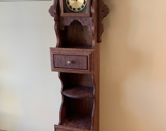 May include: A tall, dark wood grandfather clock with a decorative top, a clock face, a small drawer, shelves, and a cabinet door at the bottom.