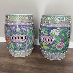 Op de afbeelding: Twee bijpassende keramische tuinbanken. De krukken zijn tonvormig met een lavendelkleurige achtergrond en bloemmotieven in roze, groen, geel en oranje. Elke kruk heeft twee opengewerkte geometrische ontwerpen en een gestreepte basis.