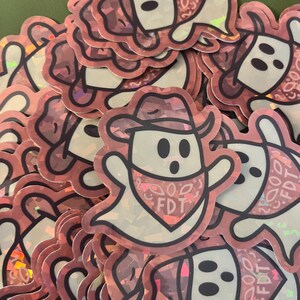 May include: A collection of holographic stickers featuring a cartoon ghost wearing a cowboy hat and a bandana. The bandana has the letters "FDT" printed on it. The stickers have a pink and white color scheme with a black outline.