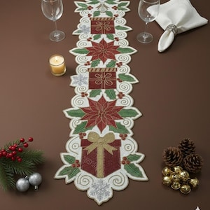 Könnte beinhalten: Ein weihnachtlicher Tischläufer mit Perlenbesatz und festlichem Design. Der Läufer zeigt rote Geschenkboxen, Weihnachtsstern und Schneeflocken mit grünen und weißen Akzenten. Ca. 152 cm lang.