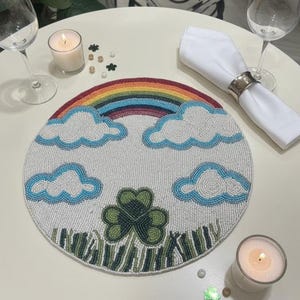 May include: A round beaded placemat featuring a rainbow, clouds, and a shamrock design. The placemat has a white background with a rainbow arching across the top. Blue and white clouds are scattered across the top. Green shamrock and grass are at the bottom.