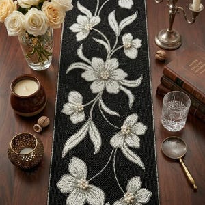 Könnte beinhalten: Schwarzer, mit Perlen besetzter Tischläufer mit einem Blumenmuster in Weiß und Creme. Der Läufer ist mit Blumen und Blättern verziert. Das Bild zeigt auch eine Vase mit Rosen, Kerzen und andere dekorative Gegenstände.