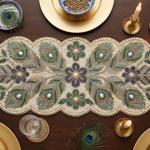 May include: A decorative beaded table runner with a peacock feather design. The runner has a cream base with green, blue, and pearl beads. The table setting includes gold plates, candles, and bowls, creating an elegant dining scene.