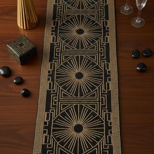 May include: A black and gold beaded table runner with an art deco design. The runner has repeating geometric patterns. A gold vase, a small box, and black stones are on the wooden surface.