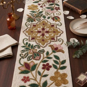 May include: Cream-colored beaded table runner with floral embroidery in gold, burgundy, pink, and green. The runner features a central medallion design and floral patterns along its length, suitable for home decor.