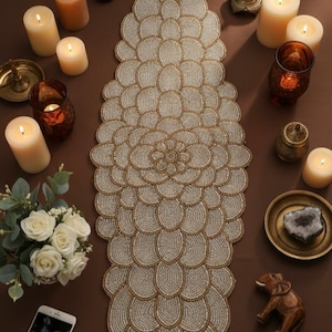 May include: A beaded table runner with a floral design, surrounded by lit candles, a bouquet of white roses, and decorative items. The runner is beige and gold, and the candles are ivory.