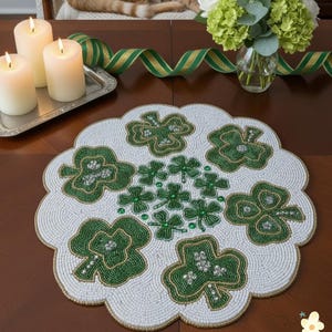 May include: A circular, white beaded placemat with a scalloped edge, decorated with green shamrock patterns. The shamrocks are adorned with green beads and silver details. A St. Patrick's Day table decoration.