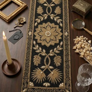 Puede incluir: Un camino de mesa negro y dorado con cuentas y un diseño floral. El camino de mesa presenta un patrón central de flores y hojas, enmarcado por un borde geométrico. Elementos decorativos como una vela, un marco y un vaso están dispuestos alrededor.