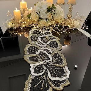 May include: A decorative table setting with a black beaded floral table runner with gold and white detailing. The centerpiece includes white flowers, candles, and gold candle holders. The table is black and reflective.
