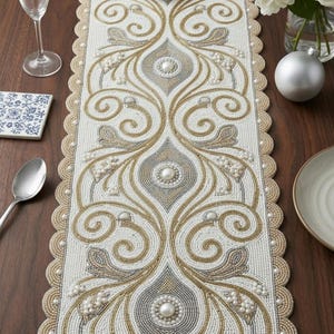 May include: A beaded table runner with an intricate design in white, gold, and gray. The runner features a scalloped edge and is adorned with pearl-like beads. The design includes swirling patterns and central medallions, suitable for home decor.