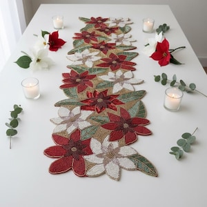 Puede incluir: Un camino de mesa con cuentas que presenta diseños de flores de pascua rojas y blancas, hojas verdes y detalles dorados. El camino de mesa está colocado sobre una mesa blanca, decorada con velas y flores.