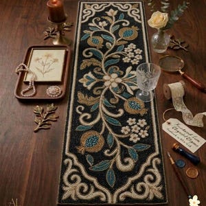 Pode incluir: Um corredor de mesa preto com contas com um design floral em dourado, verde-azulado e creme. O corredor apresenta uma videira central com romãs e flores. O corredor é cercado por itens decorativos, incluindo uma vela, uma bandeja e um copo.