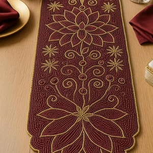 Könnte beinhalten: Burgunderfarbener, mit Perlen besetzter Tischläufer mit goldenem Blumendesign. Der Läufer zeigt große stilisierte Blumen, Sternausbrüche und Wirbelmuster. Die Kanten sind gewellt und mit goldenen Perlen verziert. Ein dekoratives Stück für einen Esstisch.