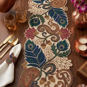 May include: A beaded table runner with a peacock feather design in blue, green, and gold. The runner is on a wooden table, with gold cutlery, glasses, and a copper pitcher. A napkin ring with a peacock design is also visible.