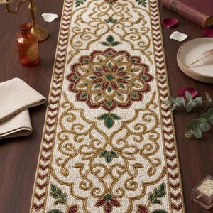 Può includere: Runner da tavolo con perline color crema con un design intricato con accenti dorati, bordeaux e verdi. Il runner ha un medaglione floreale centrale e un bordo con un motivo ripetuto. Decorativo.