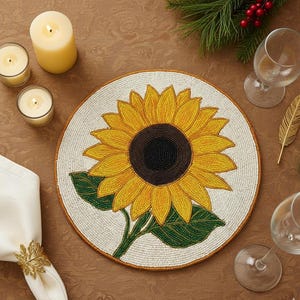 Op de afbeelding: Een ronde placemat met kralen met een zonnebloemontwerp. De placemat heeft gele bloemblaadjes en een donker centrum. De afbeelding bevat ook kaarsen, wijnglazen en een servet met een gouden ring. De diameter is ongeveer 30 cm.