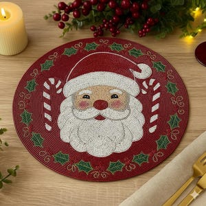 Puede incluir: Un salvamanteles navideño redondo y rojo con un diseño de Papá Noel con cuentas, con barba y gorro blancos, y dos bastones de caramelo. El borde está decorado con hojas de acebo y bayas verdes.