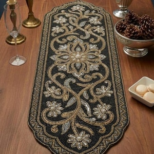 Könnte beinhalten: Schwarzer, mit Perlen bestickter Tischläufer mit floralem Design in Gold und Silber. Der Läufer ist rechteckig mit einem gewellten Rand und liegt auf einem Holztisch. Das Design zeigt eine zentrale Blume und Ranken.