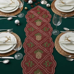 Könnte beinhalten: Festliche Weihnachtstafel mit einem bordeauxfarbenen Tischläufer mit Tannenzapfen- und Stechpalmenmotiven. Der Tisch ist mit weißen Tellern, Besteck, Weingläsern und dekorativen Serviettenringen gedeckt. Tannenzapfen, Ornamente und Kerzen vervollständigen das festliche Thema.