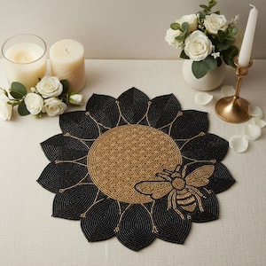 May include: A beaded sunflower-shaped placemat with a bee design. The placemat has black petals and a gold centre with a honeycomb pattern. A gold bee is on the lower right side.