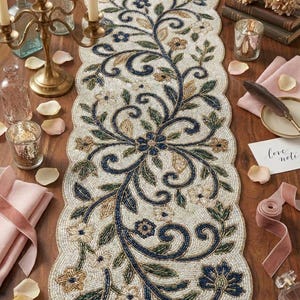 Puede incluir: Camino de mesa de cuentas color crema con un diseño floral en azul marino, verde y dorado. El camino de mesa presenta un borde festoneado y está decorado con flores y espirales. Ideal para añadir un toque de elegancia a cualquier mesa.