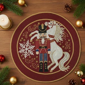 Op de afbeelding: Een ronde, bordeauxrode, met kralen bezette placemat met een gouden rand. Het ontwerp toont een notenkrakersoldaat en een wit paard. Versieringen zijn een sneeuwvlok, hulst en sterren. Kerstdecoratie.