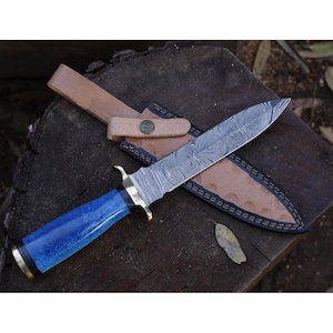 Puede incluir: Un cuchillo de caza artesanal con una hoja de acero estampada y un mango azul vibrante. El cuchillo va acompañado de una funda de cuero marrón con costuras decorativas y una trabilla para el cinturón. La hoja tiene un patrón arremolinado único.