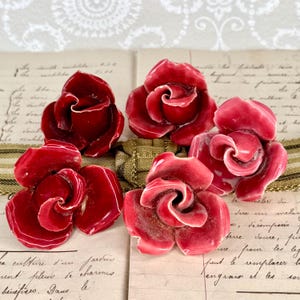 May include: Six red rose-shaped decorative objects with white accents. The roses are arranged on a vintage paper background with handwritten text and a gold and green ribbon.