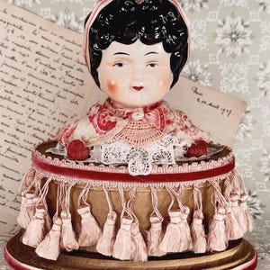 May include: A porcelain half-doll pincushion featuring a figure with dark curly hair, a pink ribbon, and a lace collar. The base is a round, gold-colored container adorned with a deep red velvet trim and a fringe of light pink tassels.