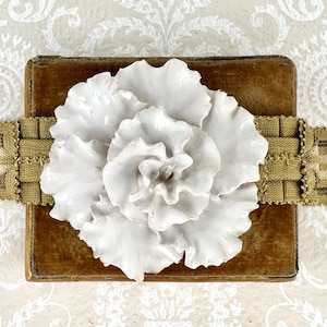 May include: A white ceramic flower is centered on a brown, rectangular object, possibly a box or book. A woven, olive-green ribbon wraps around the object. The background features a cream-colored, patterned surface.