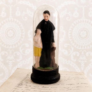 May include: A figurine of a person in a black robe standing with two smaller figures, one wearing yellow shorts and one in a brown robe, all enclosed within a clear glass dome on a circular black base, creating a decorative display piece.