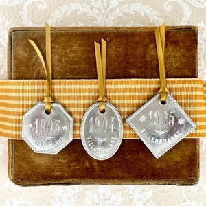 May include: Three silver-colored tags with the years 1903, 1904, and 1905, each suspended by a gold ribbon. The tags are in different shapes: an octagon, an oval, and a diamond. The tags are on a brown velvet-covered box with a striped ribbon.