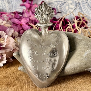 May include: A silver-toned metal heart-shaped ex-voto ornament featuring a flame at the top. The surface is engraved with the text: "Ama Mere cherie J De Raedt Conseillere 21 Avril 1889". The object has a smooth, reflective finish and a pointed base.