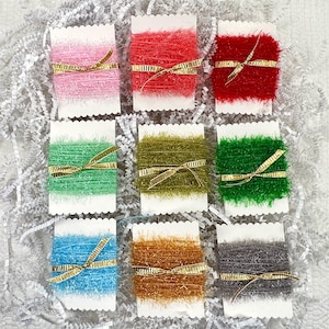 May include: Nine spools of colorful yarn, each wrapped with a gold ribbon, are displayed on white paper. The yarn colors include pink, red, green, olive, dark green, blue, orange, and gray. The yarn is fluffy and textured.