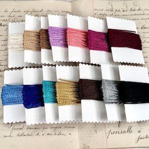 May include: Assortment of 12 spools of colorful waxed cotton cord, each wound on a white card with scalloped edges. The cords are arranged in two rows of six, with colors including beige, pink, purple, red, burgundy, blue, teal, gold, brown, gray, and black.