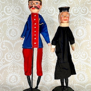May include: Two antique wooden dolls. One doll is dressed in a blue jacket with red trim and red pants. The other doll is dressed in a black dress and hat. Both dolls are standing on black circular bases.