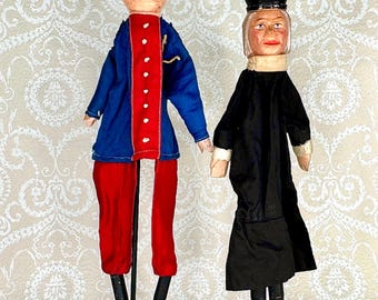 May include: Two antique wooden dolls. One doll is dressed in a blue jacket with red trim and red pants. The other doll is dressed in a black dress and hat. Both dolls are standing on black circular bases.