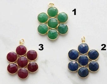 Puede incluir: Tres colgantes de piedras preciosas con forma de flor. Uno presenta piedras verdes, otro piedras azules y el tercero piedras rojas. Cada piedra está engastada en un bisel dorado con un pequeño lazo para colgar.