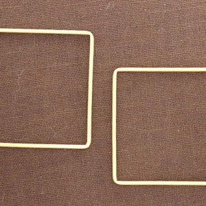 Puede incluir: Dos aros metálicos cuadrados de color amarillo pálido. Los aros están sobre un fondo de tela marrón.