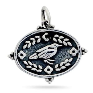 Puede incluir: Encanto de plata con un pájaro estilizado con las alas extendidas, rodeado de hojas y flores, en un marco ovalado.