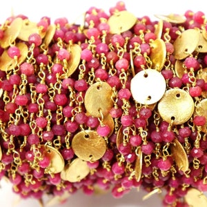 May include: A close-up of a beaded chain with small, faceted, ruby-colored beads and gold-colored, flat, round disc charms. The chain is strung on gold-colored wire.