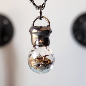 May include: A silver chain necklace with a pendant made from a clear glass bulb filled with gold-colored watch gears.