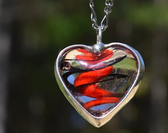 May include: A silver heart-shaped pendant necklace with a red and black swirl design inside the heart.