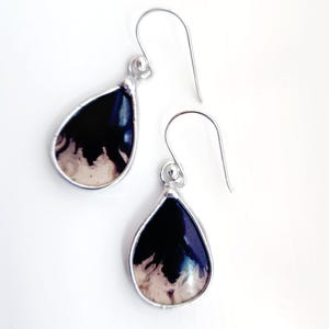 May include: A pair of teardrop-shaped earrings with a silver-colored frame. The earrings feature a black and white marbled design within the frame. The ear wires are silver-colored. The earrings are displayed on a white background.