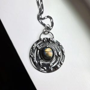 May include: A silver-toned pendant necklace with a circular design. The pendant features a central, iridescent stone surrounded by textured metal. The chain is a dark silver color. The necklace is displayed on a white surface.