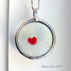 May include: A silver pendant necklace featuring a clear glass locket. Inside the locket is a small red heart and tiny clear beads. The necklace is suspended from a silver chain. The text "Bayou Glass Arts" is visible.