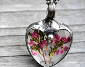 May include: A heart-shaped pendant necklace with a clear resin center, showcasing dried pink flowers. The pendant is encased in a silver-toned metal frame and hangs from a matching chain. The necklace is displayed against a gray wooden background.