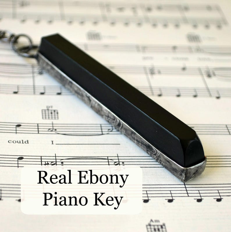 Real ebony piano key handcrafted into pendant necklace for men or women or can use as key ring, laying on a page of sheet music.